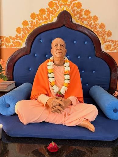 Srila Prabhupada seated, wearing a garland of flowers